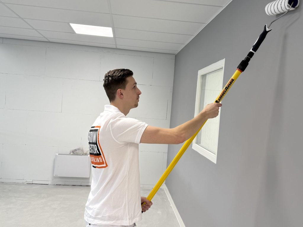 Schilder met lange roller zet een grijze sauslaag op de muur bij het raam, kalkzandsteen ernaast
